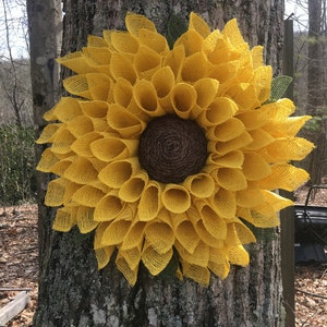 May include: A large yellow burlap sunflower wreath with a brown centre. The wreath is hanging on a tree trunk.