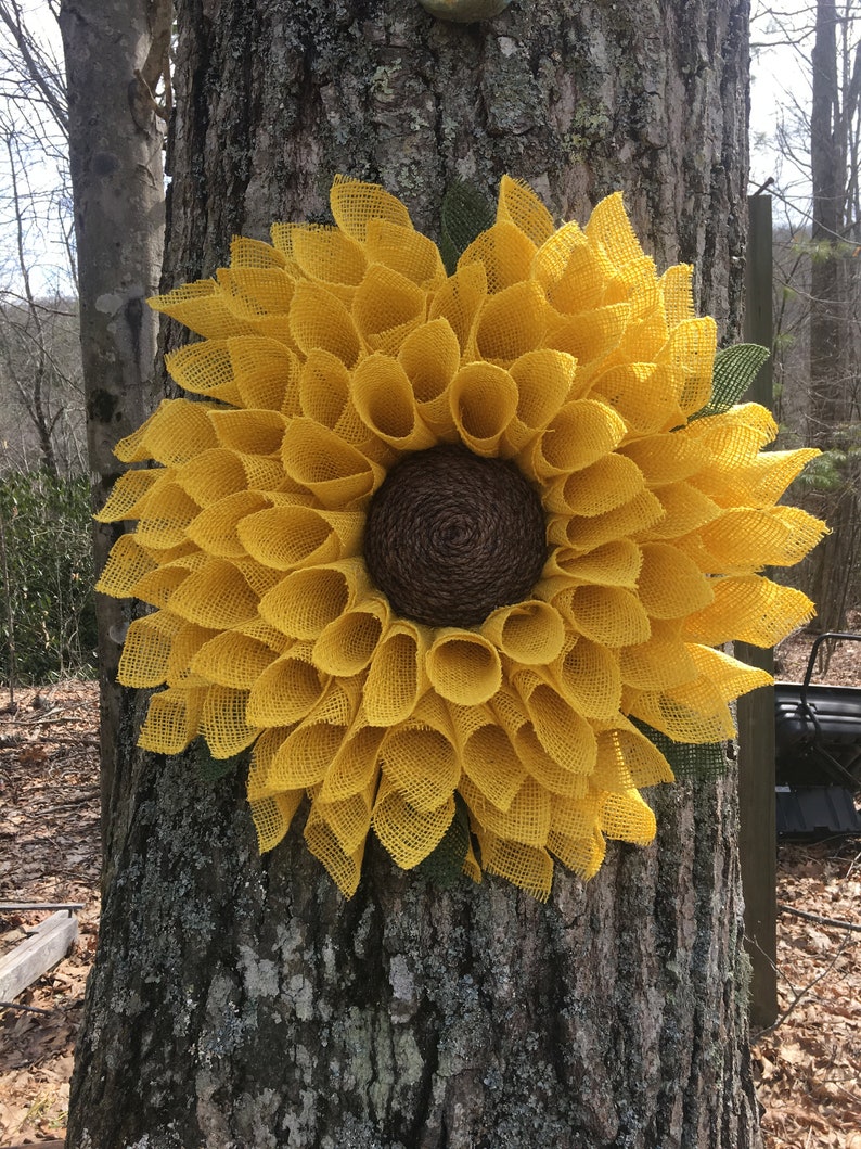 May include: A large yellow burlap sunflower wreath with a brown centre. The wreath is hanging on a tree trunk.