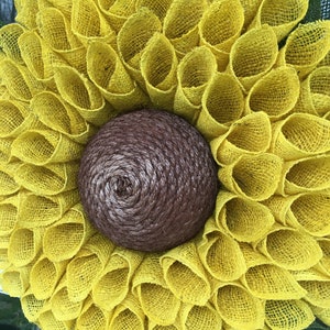 May include: A yellow burlap sunflower wreath with a brown burlap centre. The wreath is made of multiple layers of burlap, creating a full and fluffy look.