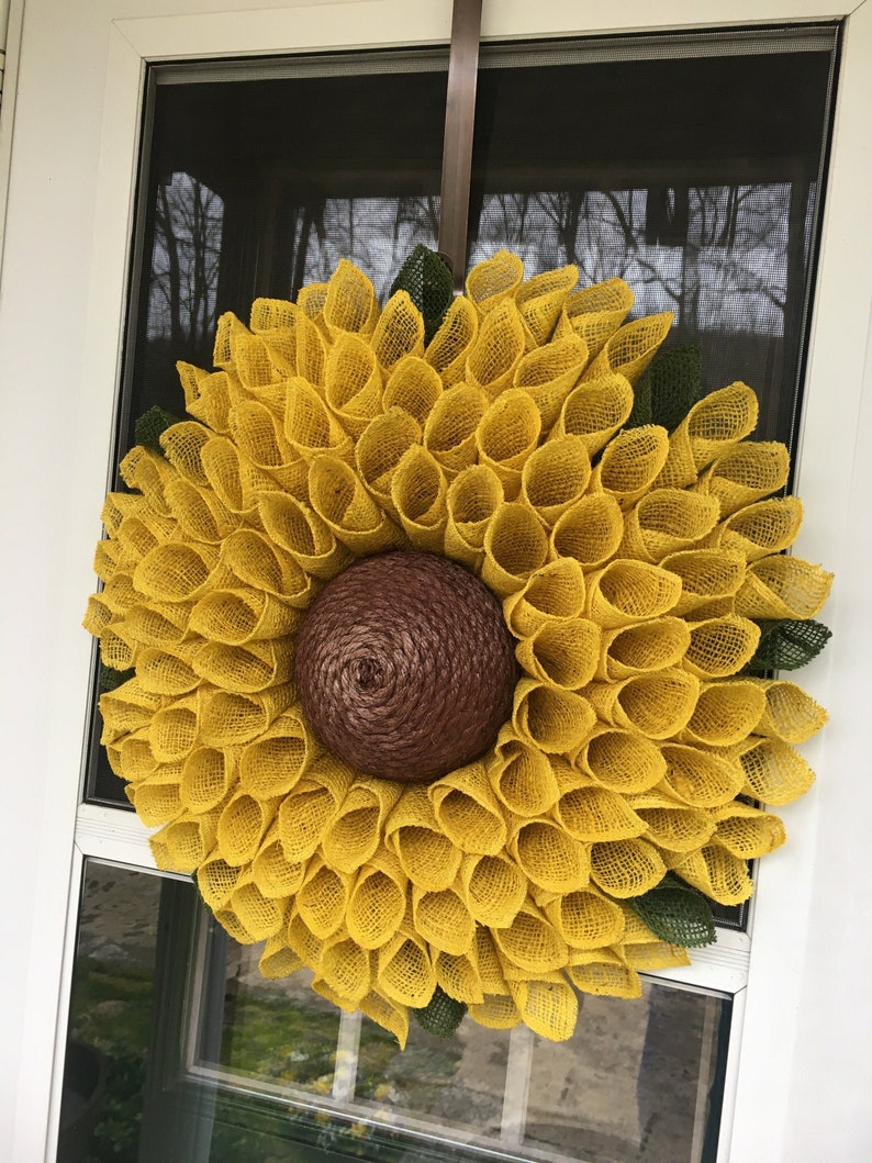 May include: A yellow burlap sunflower wreath with a brown burlap centre. The wreath is hanging on a white door.