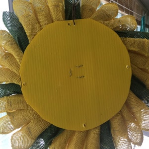 May include: A yellow and green burlap sunflower wreath with a yellow cardboard backing. The wreath is attached to the backing with wire staples.