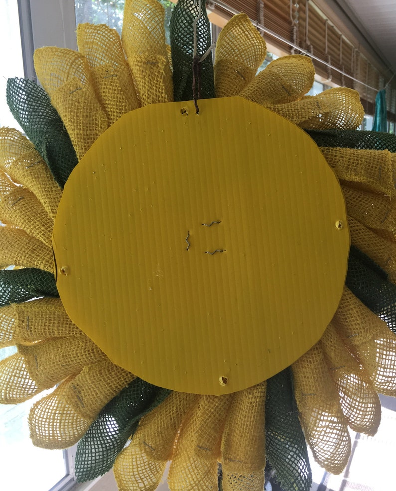 May include: A yellow and green burlap sunflower wreath with a yellow cardboard backing. The wreath is attached to the backing with wire staples.