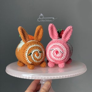 Puede incluir: Dos cake pops de conejito tejidos a crochet, uno naranja y blanco con una zanahoria en la parte superior, y otro rosa y blanco con una fresa en la parte superior. Los cake pops están sobre un soporte para pasteles blanco.