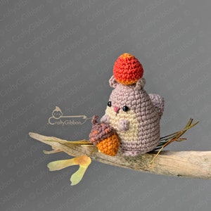 May include: A crocheted squirrel toy holding an acorn and wearing a red crocheted acorn hat. The squirrel is sitting on a branch with a leaf.