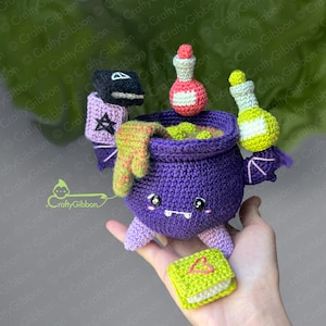 May include: A purple crocheted cauldron with a smiling face, green goo inside, and a green and pink crocheted bat wing on each side. The cauldron is holding a black book with a pentagram and a red and white potion bottle. A yellow and white potion bottle is on the other side. A green book with a heart on the cover is in the foreground.