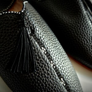 Op de afbeelding: Close-up van een paar zwarte leren pantoffels. De pantoffels hebben een textuuroppervlak, decoratieve stiksels en een zwarte kwast. De stiksels zijn wit en zwart. De pantoffels zijn waarschijnlijk voor gebruik binnenshuis.