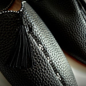 Op de afbeelding: Close-up van een paar zwarte leren pantoffels. De pantoffels hebben een textuuroppervlak, decoratieve stiksels en een zwarte kwast. De stiksels zijn wit en zwart. De pantoffels zijn waarschijnlijk voor gebruik binnenshuis.