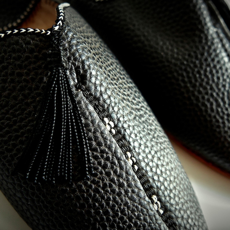 Op de afbeelding: Close-up van een paar zwarte leren pantoffels. De pantoffels hebben een textuuroppervlak, decoratieve stiksels en een zwarte kwast. De stiksels zijn wit en zwart. De pantoffels zijn waarschijnlijk voor gebruik binnenshuis.