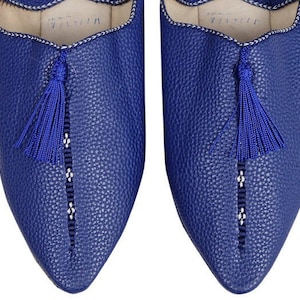 May include: A pair of blue leather slippers with a pointed toe and a tassel detail. The slippers have a white embroidered trim around the opening.