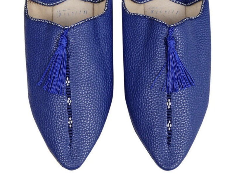 May include: A pair of blue leather slippers with a pointed toe and a tassel detail. The slippers have a white embroidered trim around the opening.