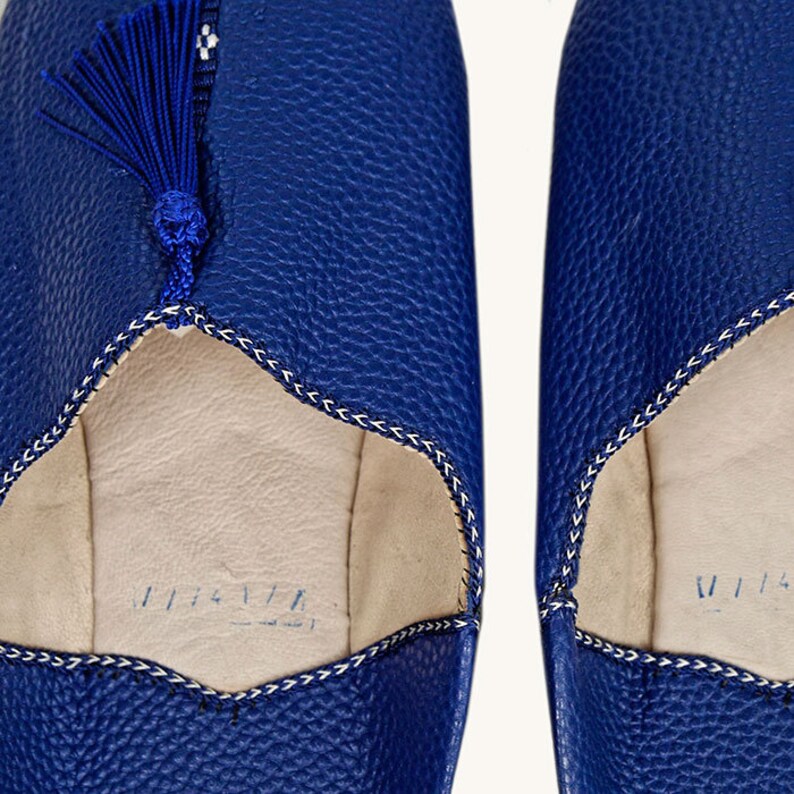 May include: A pair of blue leather slippers with a tassel detail. The slippers have a white stitching detail around the opening and a white lining.