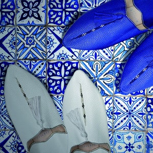 May include: Two pairs of leather slippers, one pair is white and the other is blue. The slippers are on a tiled floor with a blue and white pattern.
