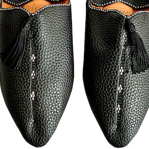 Op de afbeelding: Close-up van twee zwarte leren slippers met een getextureerd oppervlak. De slippers hebben een puntige neus, een decoratieve kwast en witte stikseldetails. De randen zijn afgewerkt met een wit en zwart patroon.