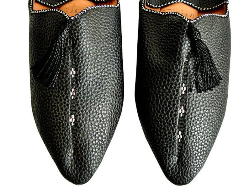 Op de afbeelding: Close-up van twee zwarte leren slippers met een getextureerd oppervlak. De slippers hebben een puntige neus, een decoratieve kwast en witte stikseldetails. De randen zijn afgewerkt met een wit en zwart patroon.