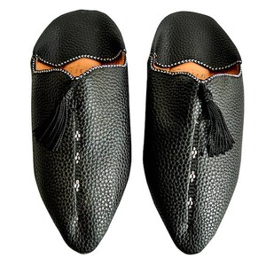 Op de afbeelding: Een paar zwarte leren slippers met een getextureerde afwerking. De slippers hebben een spitse neus, een decoratieve kwast en witte stiksels langs de naden. De binnenkant is lichtbruin.
