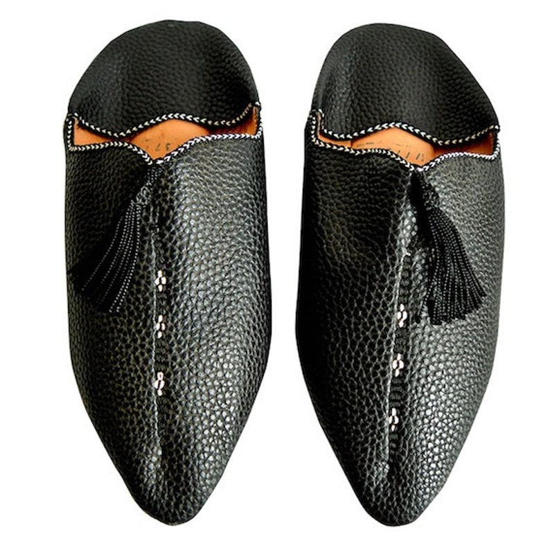 Op de afbeelding: Een paar zwarte leren slippers met een getextureerde afwerking. De slippers hebben een spitse neus, een decoratieve kwast en witte stiksels langs de naden. De binnenkant is lichtbruin.