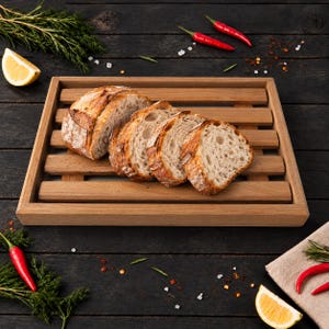 May include: A rectangular wooden bread board with a slatted design holds one loaf of crusty sourdough bread sliced into four pieces. The board is light brown, while the bread features a golden-brown crust and a porous, light-colored interior.
