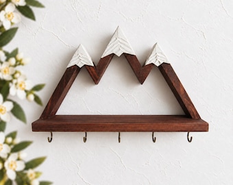 May include: A wall-mounted wooden shelf shaped like a mountain range with three peaks. The peaks are painted white to resemble snow, while the base and frame are a dark brown wood. Five small metal hooks are attached to the bottom of the shelf.