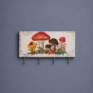 May include: A rectangular wooden wall hook rack with a distressed white finish. The front features a painted illustration of various mushrooms in red, yellow, and brown, accented with green foliage and small red berries. Four metal hooks are attached.