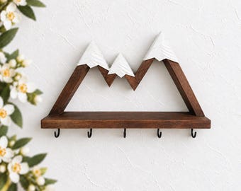 May include: A wall-mounted wooden shelf shaped like a mountain range with three peaks. The peaks are painted white with visible wood grain texture, while the base and frame are dark brown. Five black metal hooks are attached to the bottom.