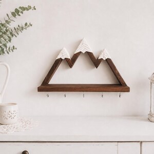 May include: A wall-mounted wooden shelf shaped like a mountain range with three peaks. The shelf is dark brown with white painted tips on the peaks and features five small silver metal hooks attached to the bottom edge for hanging items.
