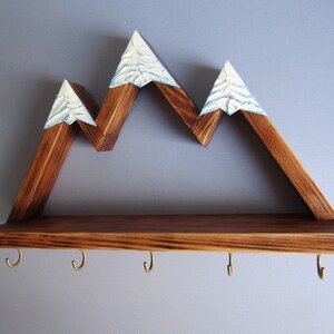 May include: Wooden mountain-shaped shelf with a brown stain and white painted snow peaks. The shelf has a lower shelf with five gold hooks.