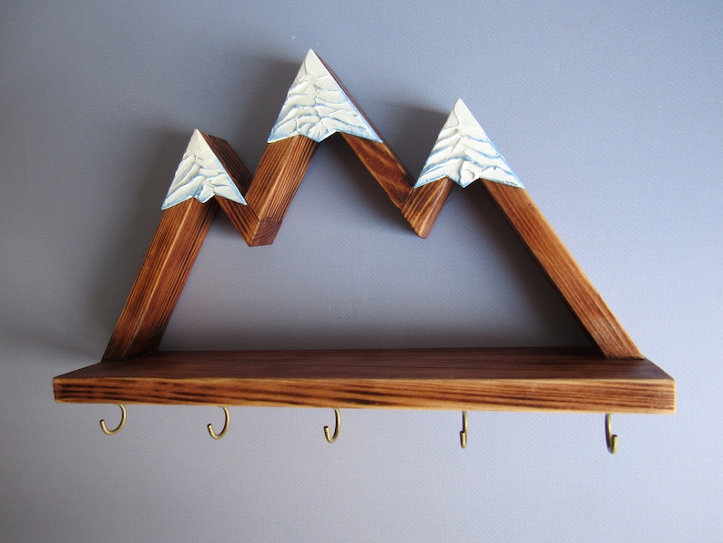 May include: Wooden mountain-shaped shelf with a brown stain and white painted snow peaks. The shelf has a lower shelf with five gold hooks.