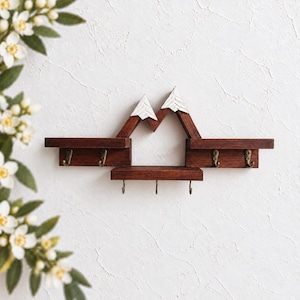 May include: A wall-mounted wooden shelf featuring a mountain-shaped design with white-painted peaks and dark brown wood grain. The unit includes two upper side shelves and one lower central shelf, equipped with a total of seven bronze-toned metal hooks.