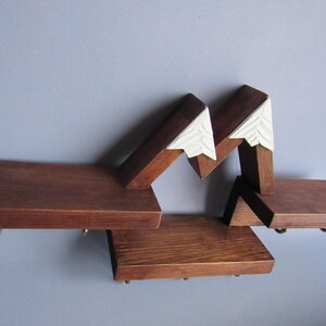 May include: A wall-mounted wooden shelf featuring three horizontal platforms connected by angled supports. The upper sections are topped with white, triangular, carved accents that resemble snow-capped mountain peaks. Small metal hooks are attached.