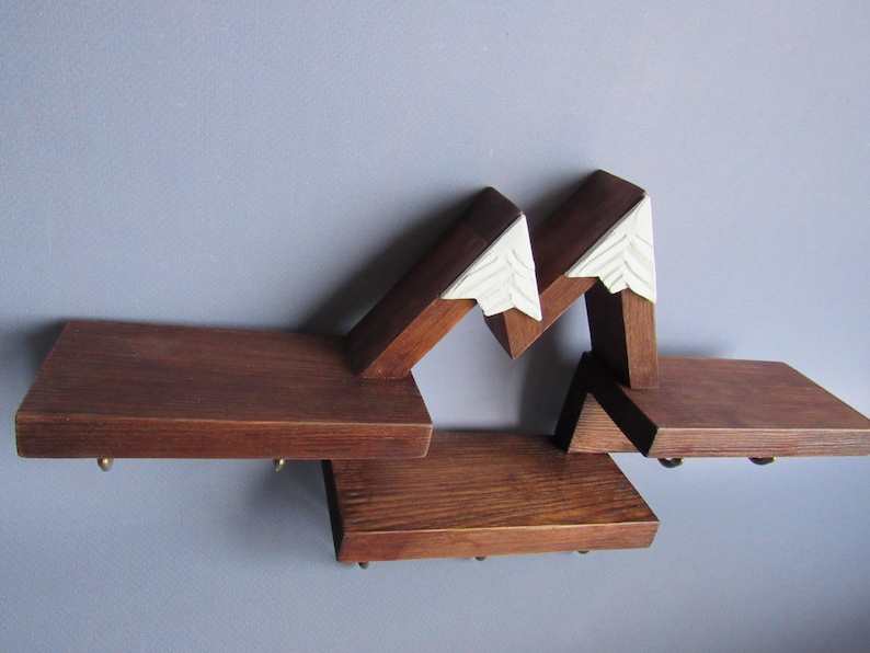 May include: A wall-mounted wooden shelf featuring three horizontal platforms connected by angled supports. The upper sections are topped with white, triangular, carved accents that resemble snow-capped mountain peaks. Small metal hooks are attached.
