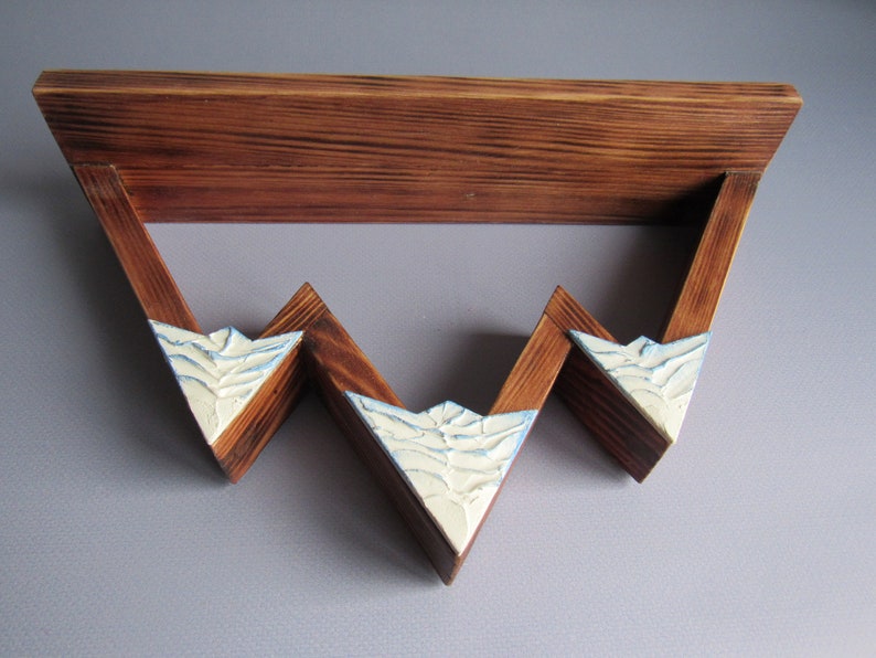 May include: A wooden shelf with a triangular design. The shelf has three tiers and is painted in a light brown color. The front of each tier is painted white with a textured design.