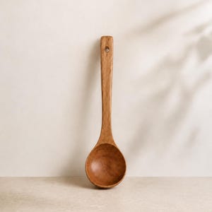 Könnte beinhalten: Eine mittelbraune Holzschöpfkelle mit einer glatten, abgerundeten Laffe und einem langen, geraden Stiel. Der Stiel verfügt am oberen Ende über ein kleines, kreisförmiges Loch zum Aufhängen. Die Holzmaserung ist auf der Oberfläche sichtbar.