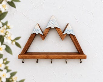 May include: A wall-mounted wooden shelf shaped like a mountain range with three peaks. The peaks feature painted white and light blue snow-capped details. The base of the shelf includes five dark metal hooks for hanging items.