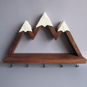 May include: A wall-mounted shelf crafted from dark brown stained wood, featuring a mountain range silhouette with three peaks topped in white paint. The horizontal base includes five small metal hooks attached to the underside for hanging items.