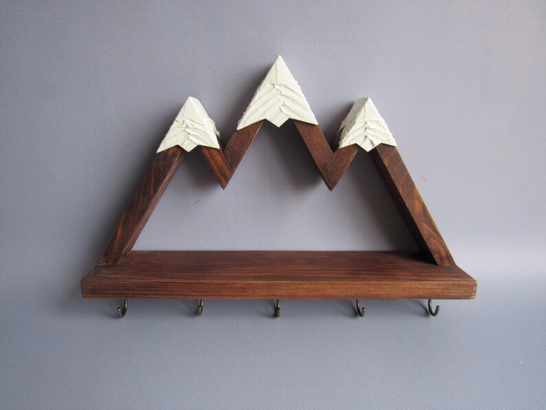 May include: A wall-mounted shelf crafted from dark brown stained wood, featuring a mountain range silhouette with three peaks topped in white paint. The horizontal base includes five small metal hooks attached to the underside for hanging items.
