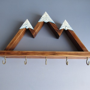 May include: Wooden mountain-shaped shelf with five hooks underneath. The shelf is stained brown and has white painted peaks.
