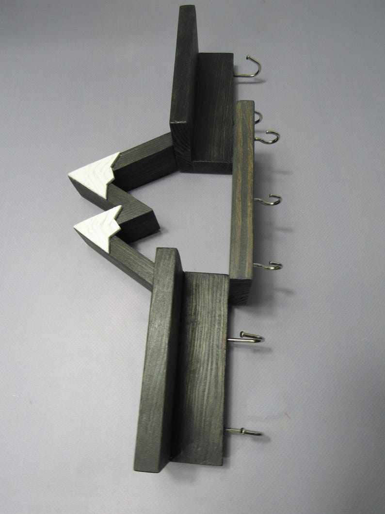May include: A wall-mounted wooden organizer featuring a dark charcoal finish with two white-painted triangular mountain peaks. The structure includes multiple horizontal shelves and five silver-toned metal hooks for hanging items in a vertical arrangement.