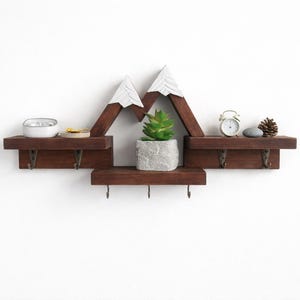 May include: A dark brown wooden wall shelf shaped like mountain peaks with white painted tops. The shelf features three horizontal tiers and seven small metal hooks attached to the bottom edges for hanging items, displaying a small plant.