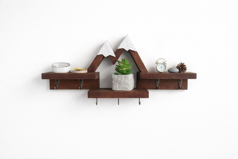 May include: A dark brown wooden wall shelf shaped like mountain peaks with white painted tops. The shelf features three horizontal tiers and seven small metal hooks attached to the bottom edges for hanging items, displaying a small plant.