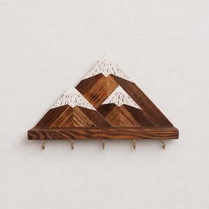 Könnte beinhalten: Wandregal aus Holz in Form einer Bergkette, dunkelbraun lackiert. Das Design zeigt drei Berggipfel mit weißen Akzenten und vier Messing-Haken. Ein dekoratives und praktisches Element für die Wand.