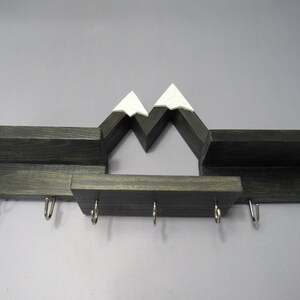 May include: A dark wood wall-mounted shelf featuring a mountain range silhouette with white-painted peaks. The unit includes three horizontal display surfaces and seven silver-colored metal hooks attached to the bottom edge for hanging items.