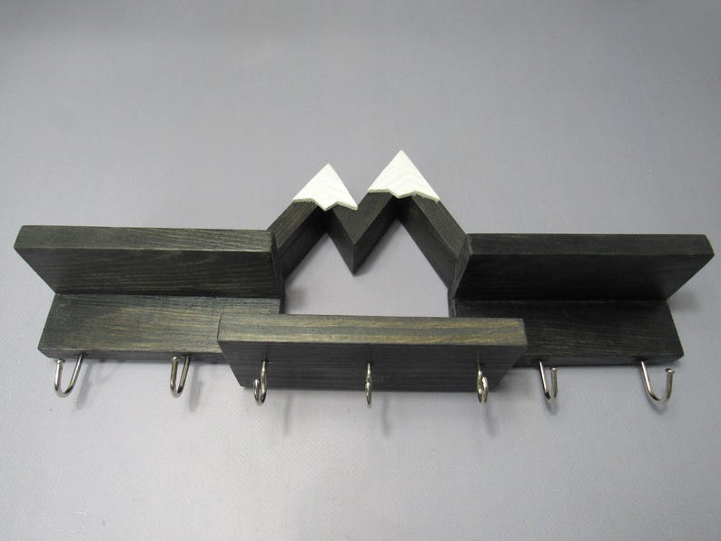 May include: A dark wood wall-mounted shelf featuring a mountain range silhouette with white-painted peaks. The unit includes three horizontal display surfaces and seven silver-colored metal hooks attached to the bottom edge for hanging items.