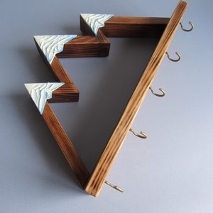 May include: Wooden wall-mounted key holder with a mountain range design. The holder has five hooks and is painted in a dark brown color with white accents.