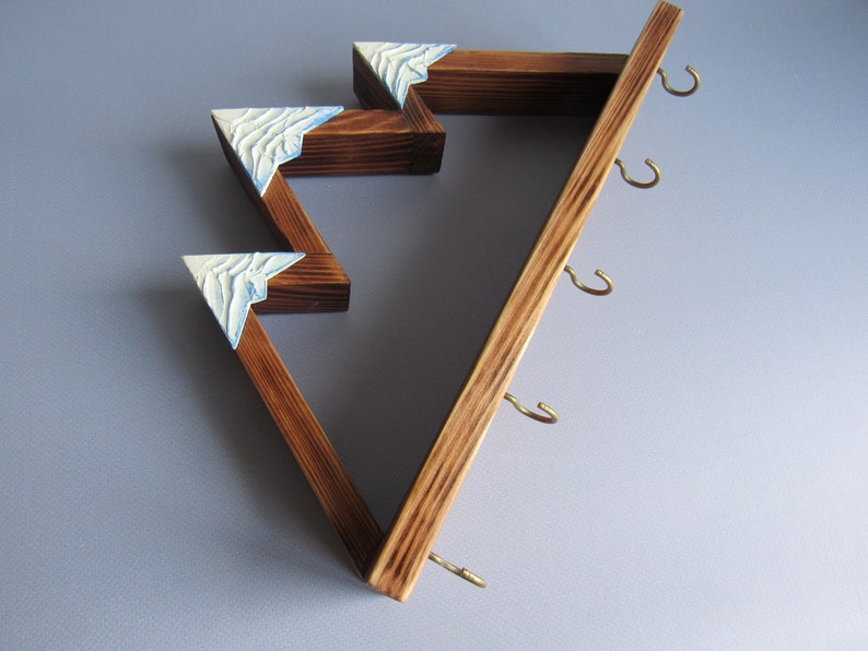 May include: Wooden wall-mounted key holder with a mountain range design. The holder has five hooks and is painted in a dark brown color with white accents.