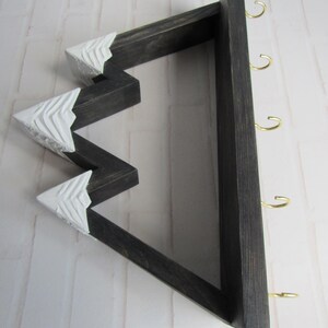 May include: A wall-mounted wooden key holder shaped like a mountain range with a dark stained finish. The three peaks are painted white with carved texture details. The flat base features four gold-colored metal hooks for hanging items.