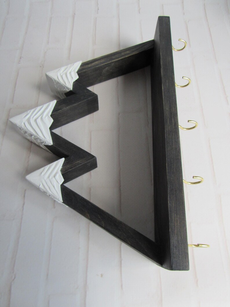 May include: A wall-mounted wooden key holder shaped like a mountain range with a dark stained finish. The three peaks are painted white with carved texture details. The flat base features four gold-colored metal hooks for hanging items.