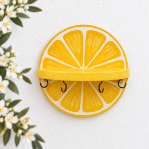 May include: A circular wall-mounted shelf shaped like a sliced lemon, featuring a bright yellow and white citrus segment pattern. The shelf includes a horizontal ledge and four dark metal hooks attached to the bottom edge for hanging items.