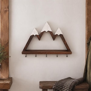 May include: A wall-mounted wooden shelf shaped like a mountain range with three peaks. The peaks are painted white to resemble snow, while the base and frame are dark brown wood. The shelf features five small metal hooks attached to the bottom edge.