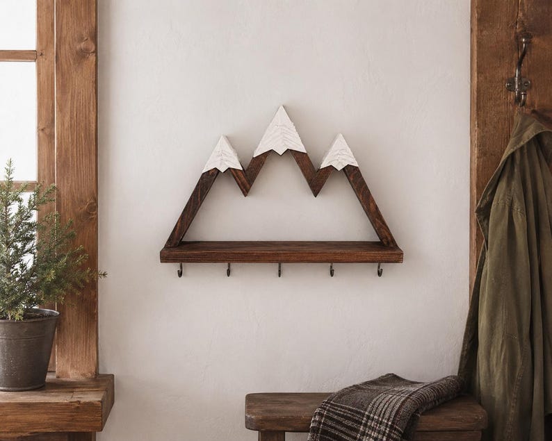 May include: A wall-mounted wooden shelf shaped like a mountain range with three peaks. The peaks are painted white to resemble snow, while the base and frame are dark brown wood. The shelf features five small metal hooks attached to the bottom edge.