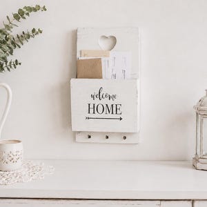 May include: A white wooden wall-mounted mail organiser featuring a heart-shaped cutout at the top, the black text "welcome HOME" with an arrow graphic, and three small metal hooks at the bottom for hanging items, holding several envelopes.
